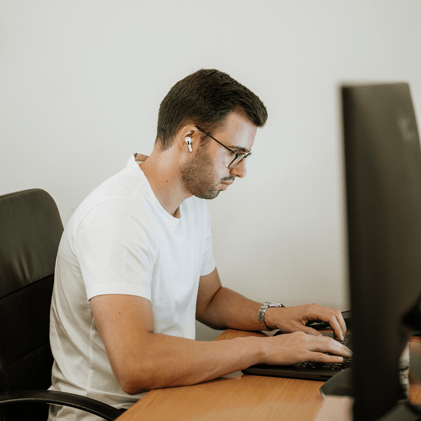 João Reis working on his computer at the Mediaweb's office, wearing a white t-shirt, glasses and hearpods on